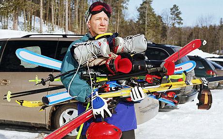 Male skier in a car park struggling to carry skis, poles, boots etc for the whole family
