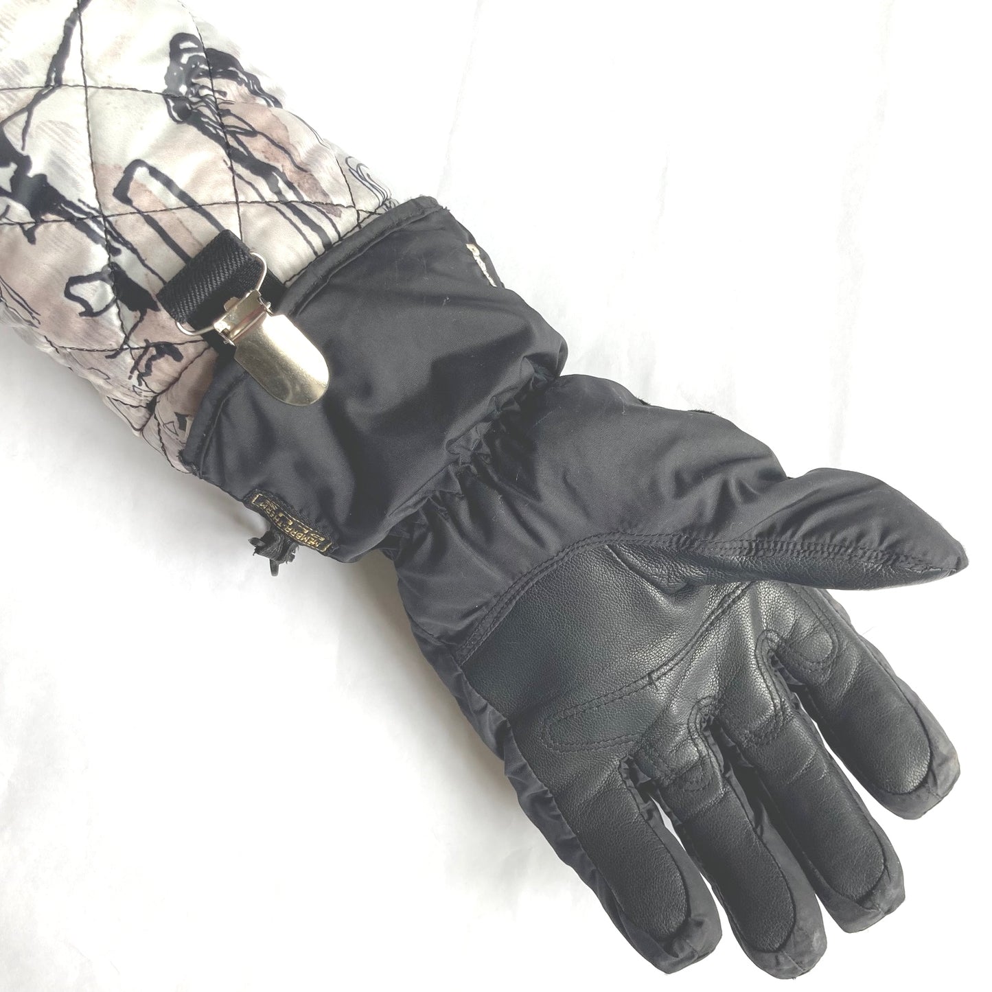 Close up of a black Clip2Cuff attached to a cuff at one end and a glove at the other end of the elastic strap