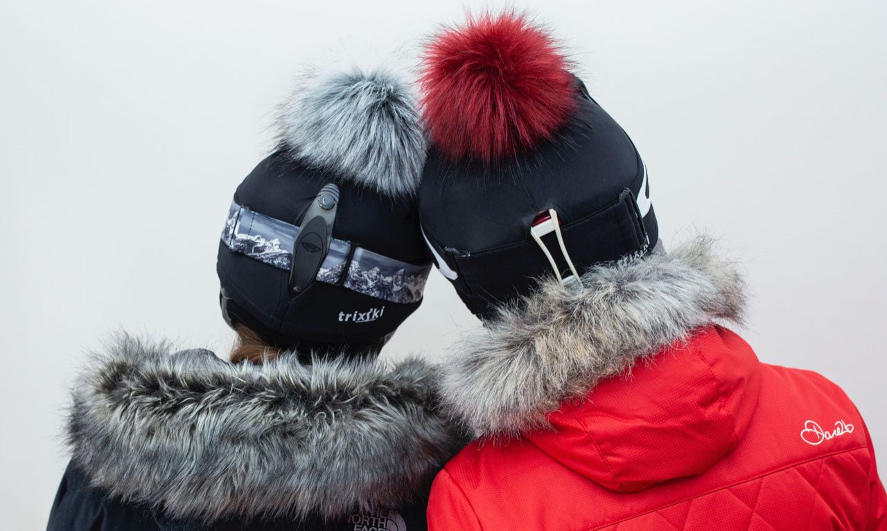 Back view of two skiers, each wearing a trixski Ski Helmet Cover.  One has red and the other has grey Faux Fur Pom Pom